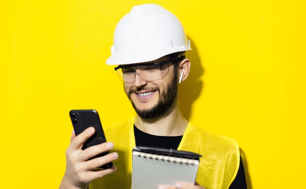 Young smiling man architect, builder engineer, wearing white construction safety helmet, glasses and jacket. Using smartphone and wireless earphones, isolated on yellow wall.