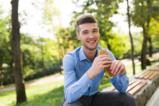 Young smiling man in blue shirt sitting on bench with sandwich in hands joyfully looking in camera while spending time in cozy green park