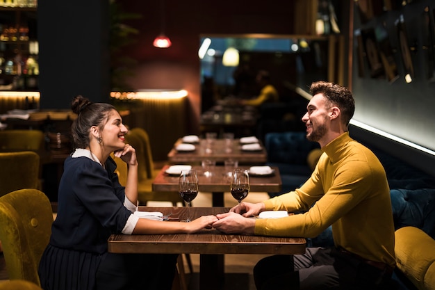 Giovane uomo sorridente e donna allegra che si tengono per mano al tavolo con bicchieri di vino nel ristorante