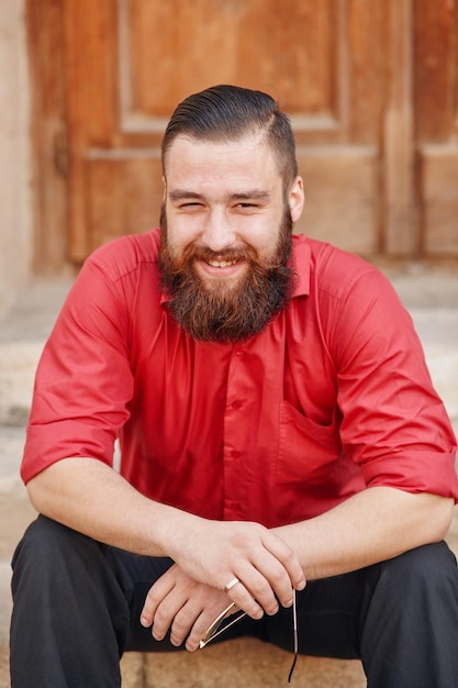 Young smiling man dressed in red shirt and black trousers sitting on the porch