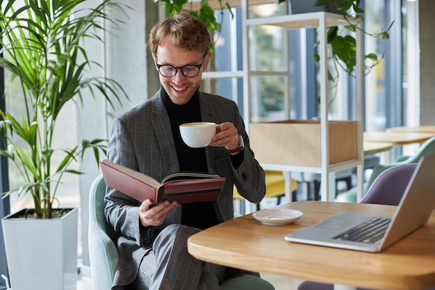 Young smiling man holding cup of coffee reading book in cafe. Coffee break concept