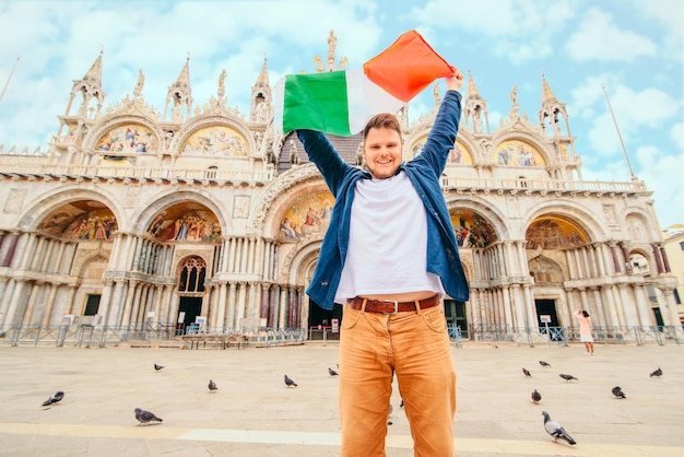 Photo young smiling man holding italian flag above his head in font of saint marks basilica