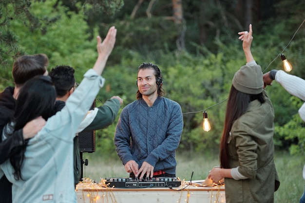 Young smiling man in jacket and headphones making music in front of crowd of his friends dancing and enjoying summer weekend