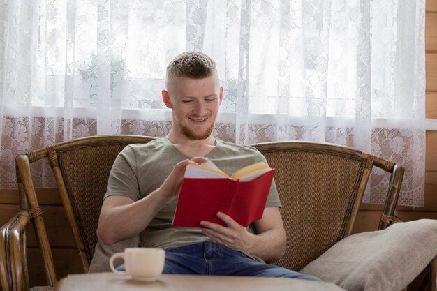 Young smiling man reading book with red cover on wicker bench in countryside wooden house