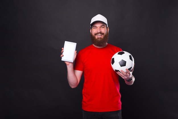 молодой улыбающийся человек показывает планшет и держит футбольный мяч над черным