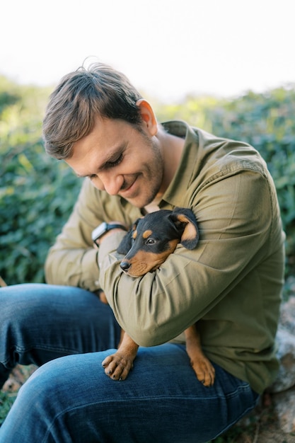 Young smiling man sitting with a puppy on his knees hugging and looking at him