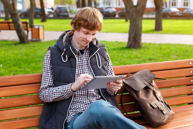 Young smiling man with a backpack in the park reading a social networks via tablet