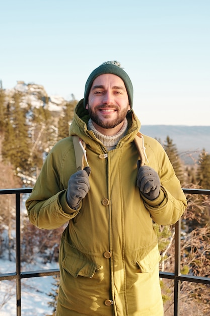 Молодой улыбающийся человек с рюкзаком стоит перед камерой на фоне деревьев и скал, покрытых снегом, и ясного голубого неба над зимним днем