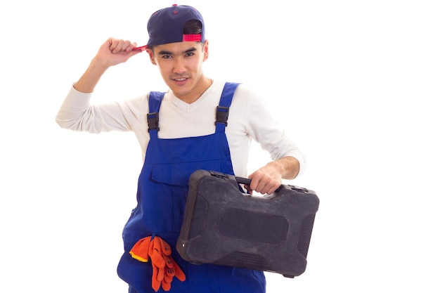 Young smiling man with black hair in white shirt and blue overall with orange gloves and blue snapback holding toolbox on white background in studio