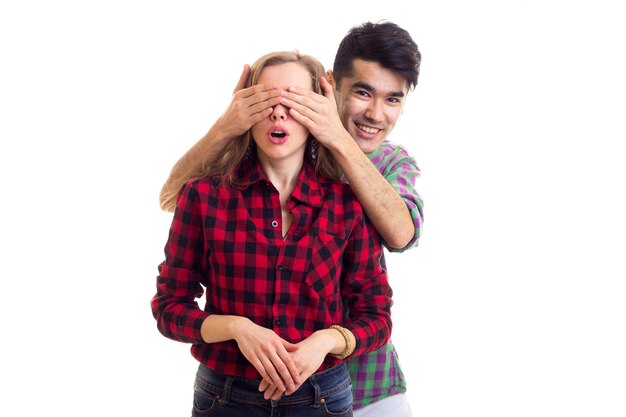 Young smiling man with dark hair closing eyes of young woman with long chestnut hair in plaid shirt