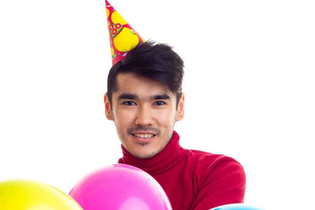 Giovane uomo sorridente con i capelli scuri in camicia rossa con cappello celebrativo che tiene molti palloncini colorati