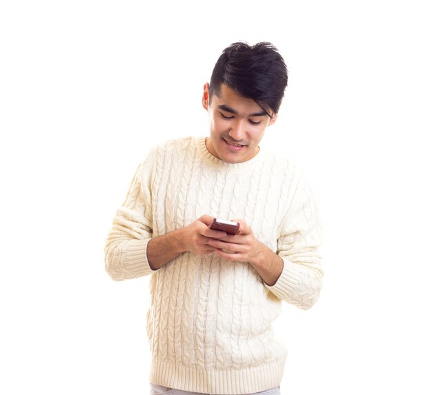 Young smiling man with dark hair in white sweater and jeans using smartphone in studio