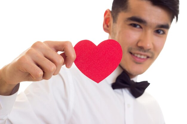 Young smiling man with dark hair in white Tshirt with black bowtie holding a red paper heart