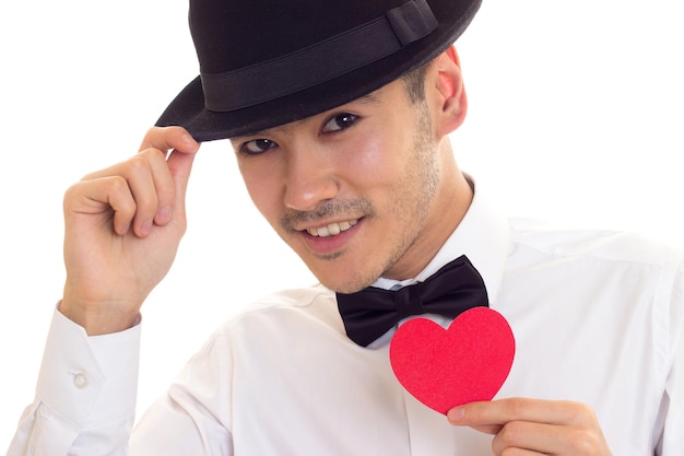 Молодой улыбающийся мужчина с темными волосами в белой футболке с бабочкой и черной шляпе держит красное бумажное сердце