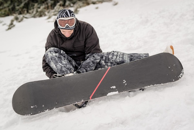Young smiling man with snowboard adjusting bindings on boot.
