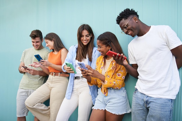 young smiling multicultural group of friends using cell phones students leaning against blue wall
