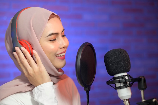 A young smiling muslim female singer wearing headphones with a microphone while recording song in a music studio with colorful lights.