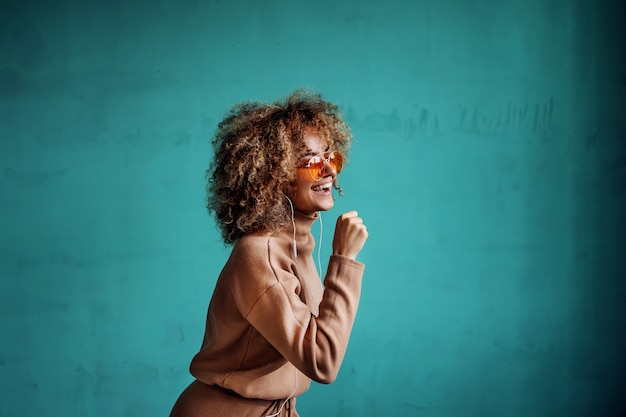 Giovane donna alla moda giocosa sorridente con i capelli ricci ascoltando la musica e il canto