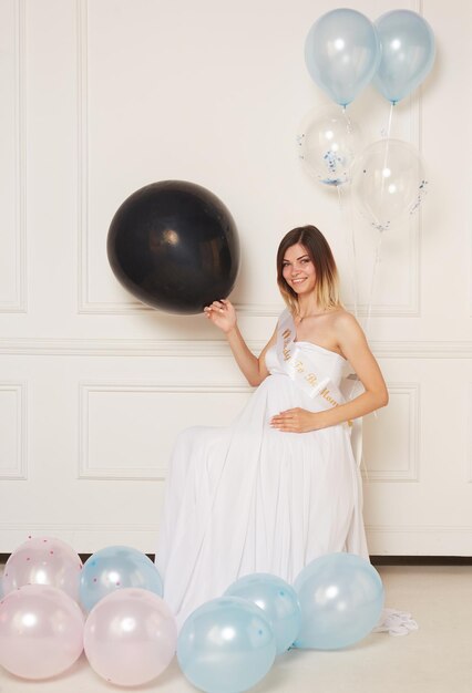 A young smiling pregnant woman in anticipation of a baby in a white dress hugs belly isolated white neoclassical wall