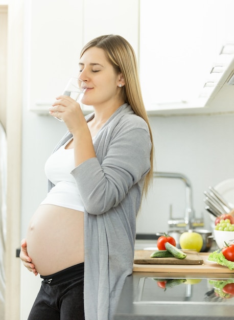 Giovane donna incinta sorridente che beve acqua in cucina?