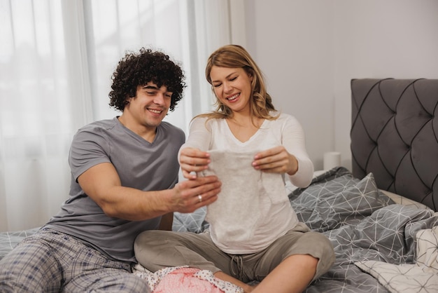 Young smiling pregnant woman showing her husband baby clothes while relaxing in bedroom.