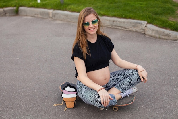 Young smiling pregnant woman with cakebag sitting on the skateboard on the background of green lawn