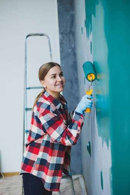 Giovane donna graziosa sorridente in camicia tenendo il rullo nella mano destra e parete di pittura riparazione nella stanza
