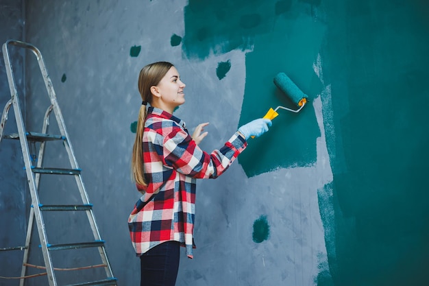 Young smiling pretty woman in shirt holding roller in right
hand and painting wall repair in the room