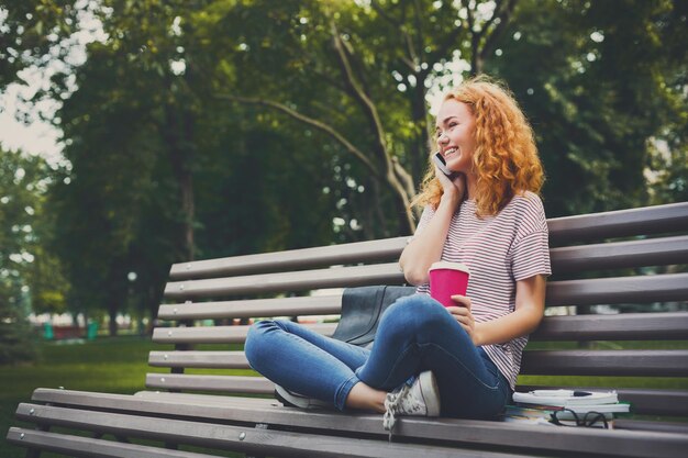 携帯電話で話し、公園のベンチに座ってコーヒーを飲む若い笑顔の赤毛の女の子。教育の概念