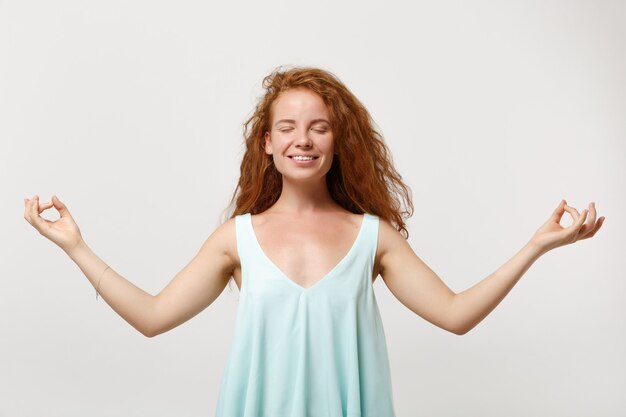 Giovane donna sorridente di redhead in vestiti casuali che posano isolati su fondo bianco. concetto di stile di vita della gente. mock up copia spazio. tieni le mani nel gesto dello yoga, rilassati meditando, tenendo gli occhi chiusi.