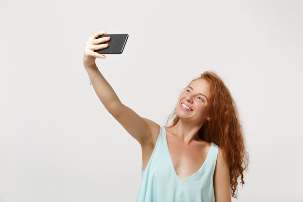 Молодая улыбающаяся рыжая девушка женщина в повседневной легкой одежде позирует изолированной на белом фоне стены, студийный портрет. Концепция образа жизни людей. Копируйте пространство для копирования. Делать селфи на мобильный телефон.