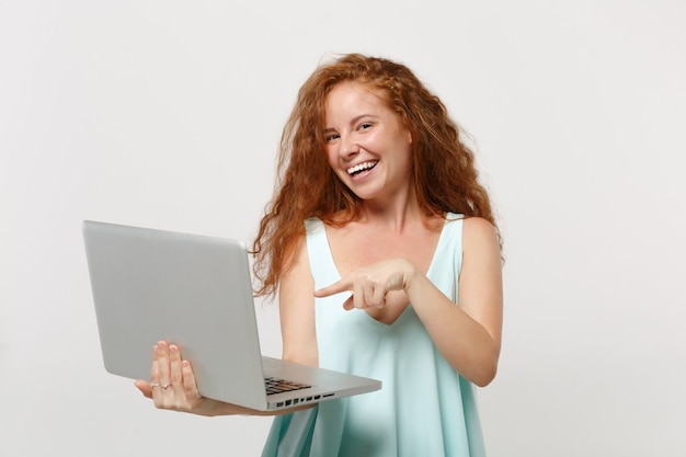 Молодая улыбающаяся рыжая девушка женщина в повседневной легкой одежде позирует изолированной на белом фоне стены. концепция образа жизни людей. копируйте пространство для копирования. работает, указывая указательным пальцем на портативный компьютер.