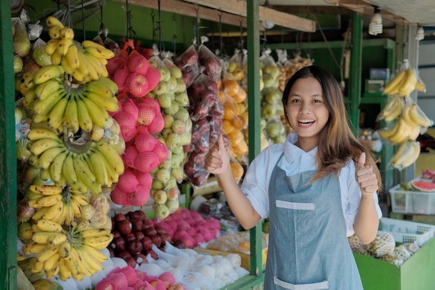 Young smiling seller in apron happily with thumbs up in fruits stall