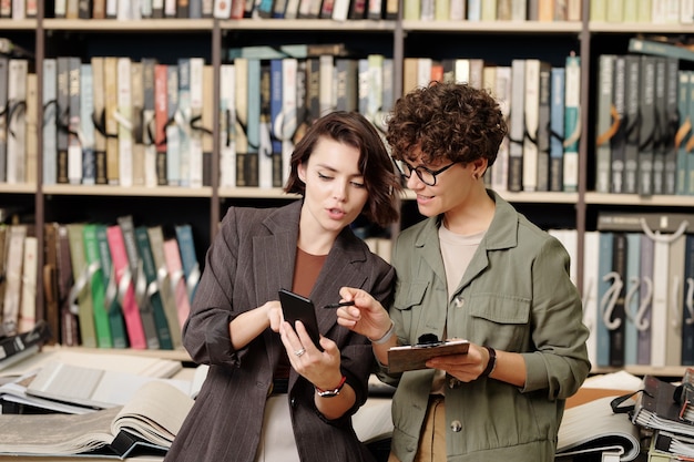 Photo young smiling shop assistant or consultant of design studio pointing at screen of smartphone held by female client scrolling through samples