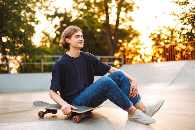 Young smiling skater in black tshirt and jeans happily spending
time with skateboard at modern skate park with sunset on
background