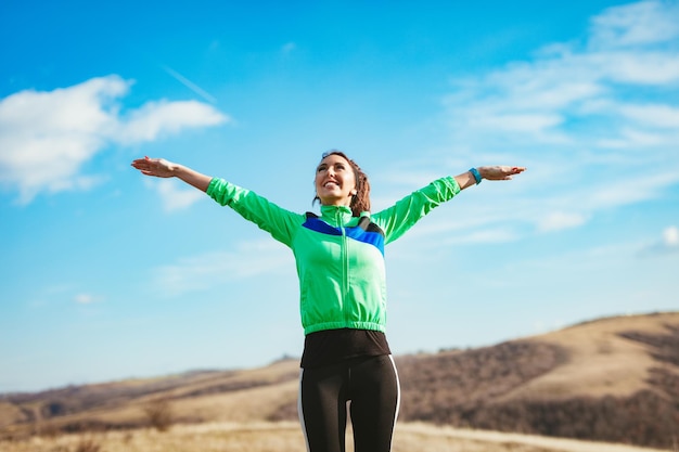 Молодая улыбающаяся спортсменка с поднятыми руками празднует успех и расслабляется после пробежки на природе. За ним ясное небо с облаками.