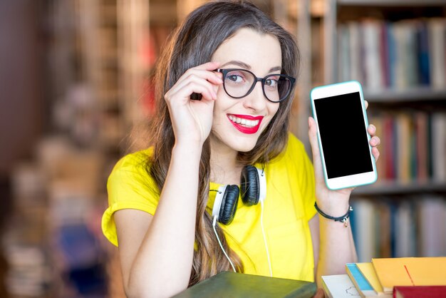 Young smiling student showing smart phone with empty screen standing at the library. Mobile application concept