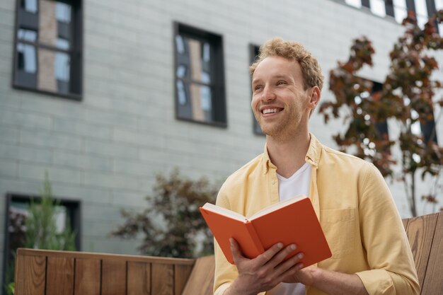 Giovane studente sorridente che studia libro di lettura seduto su una panchina nel campus universitario