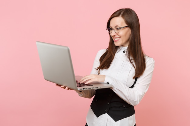 Young smiling successful business woman in glasses holding and using laptop pc computer isolated on pastel pink background. Lady boss. Achievement career wealth concept. Copy space for advertisement.