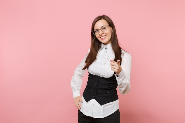 Giovane donna d'affari di successo sorridente in tuta, occhiali che puntano il dito indice sulla telecamera isolata su sfondo rosa pastello. signora capo. concetto di ricchezza di carriera di successo. copia spazio per la pubblicità.