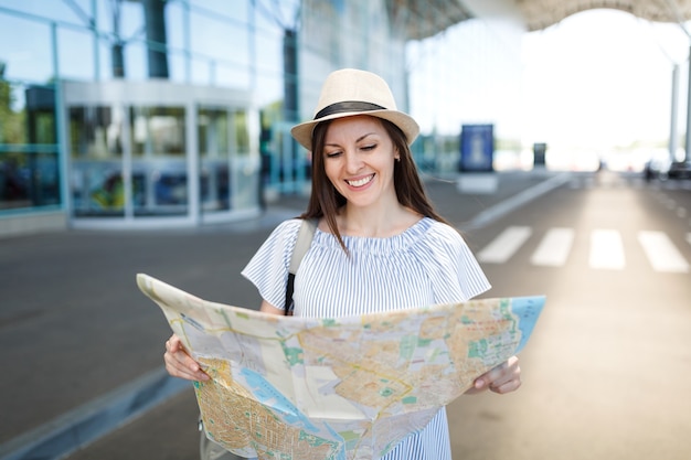 Giovane donna turistica sorridente del viaggiatore in cappello e vestiti leggeri che tengono mappa cartacea, in piedi all'aeroporto internazionale