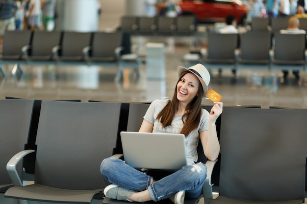 Young smiling traveler tourist woman in hat sit with crossed legs, working on laptop hold credit card wait in lobby hall at airport