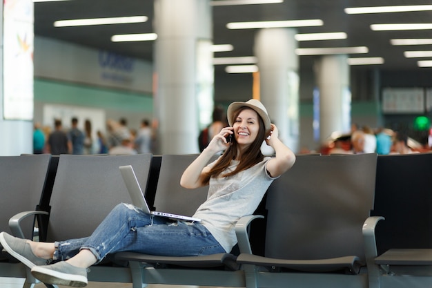 Young smiling traveler tourist woman working on laptop, talk on mobile phone, calls friend at airport