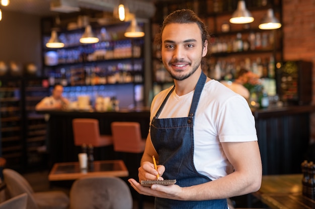 Young smiling waiter in workwear making notes in notepad while standing in front of camera on background of bar counter and interior