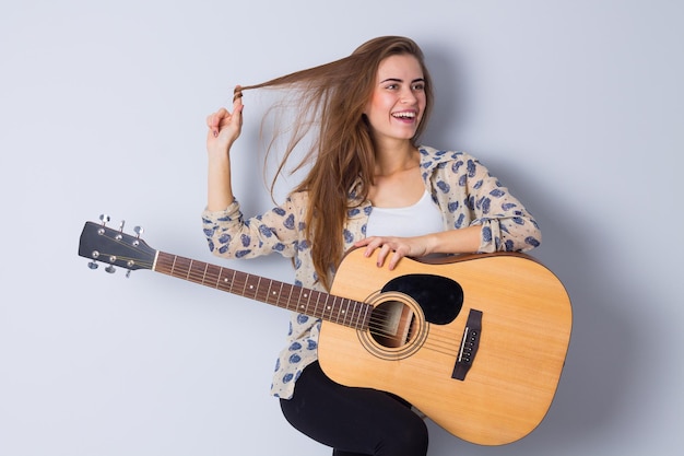 Young smiling woman in beige blouse holding a guitar on gray background in studio