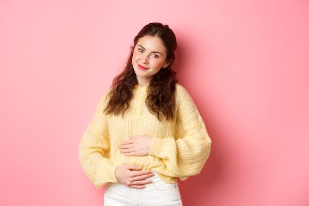 Young smiling woman being pregnant, rubbing her belly and looking with hopeful happy face at camera, expecting for baby, standing against pink wall