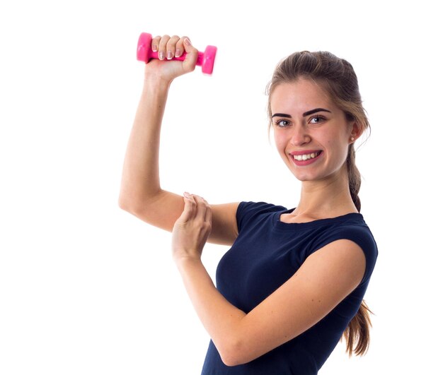 Young smiling woman in blue T-shirt holding pink dumbbell and touching her biceps on white background in studio