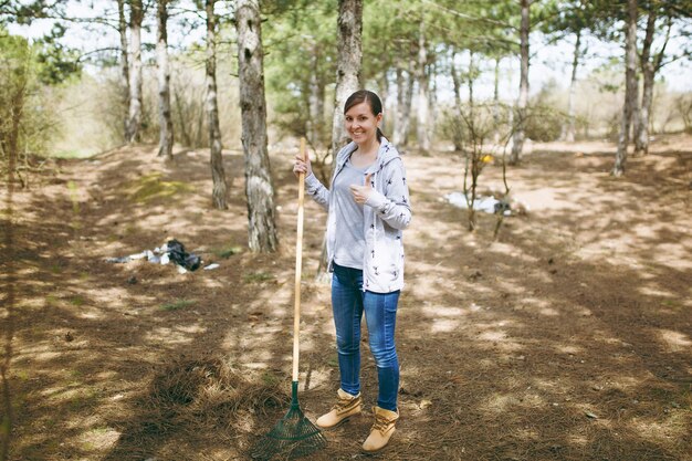 Young smiling woman cleaning using rake for garbage collection and showing thumb up in littered park. Problem of environmental pollution