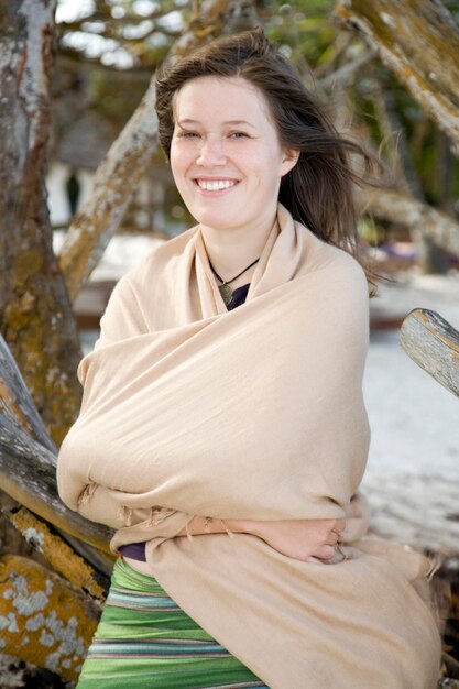 Foto giovane donna sorridente coperta da una coperta appoggiata su un tronco in spiaggia