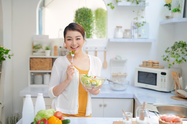 Young smiling woman eating vegetables salad at kitchen